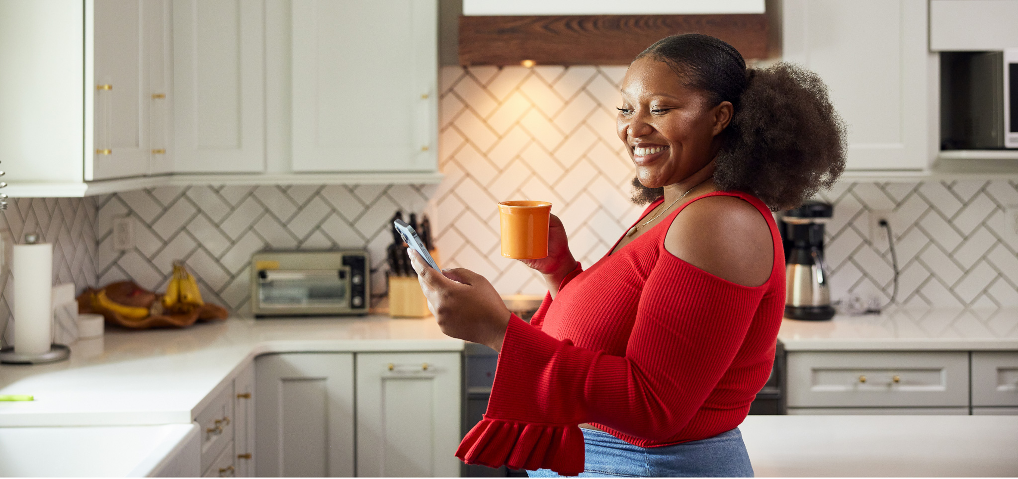 Female adult in her kitchen holding a cup of coffee in one hand and her phone in the other.