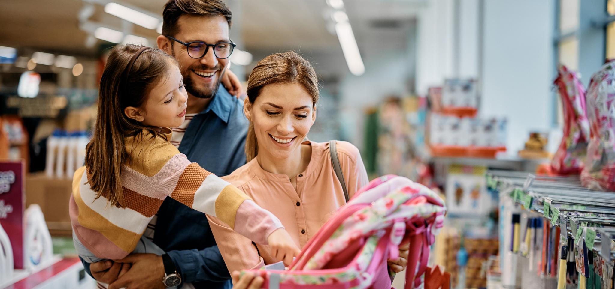 Happy little girl pointing to a backpack while buying school supplies with her parents in a store.